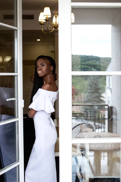 Ziemlich schlanke afrikanisch-amerikanische Frauen in einem passenden weißen Kleid posieren in der Nähe des französischen Fensters. — Stockfoto