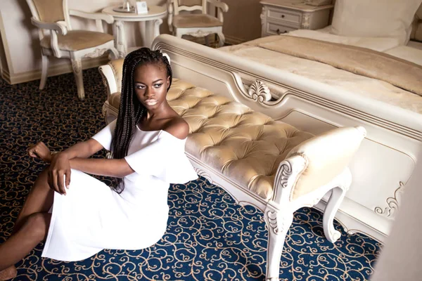 Bella ragazza afroamericana in abito bianco e con dreadlocks o trecce africane sulla testa seduta sul pavimento accanto al letto . — Foto Stock