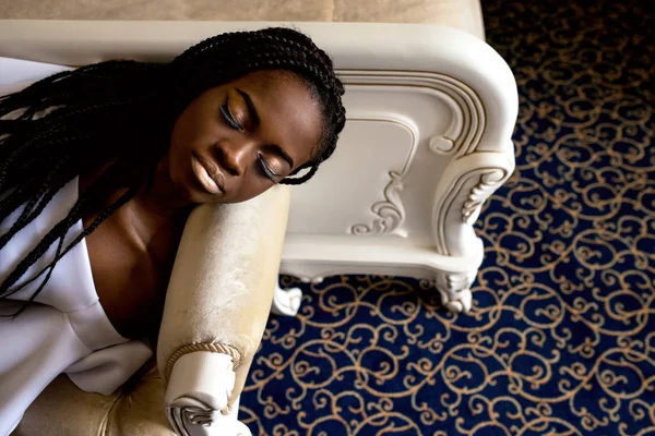 Docela africký americký žena ležící na pohovce nedaleko velké posteli ve světlé místnosti. Dívka s dredy nebo africké copánky — Stock fotografie