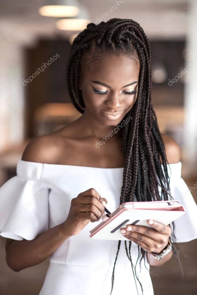 Portrait of afroamerican woman with beautiful skin tone holding the tablet in her hands. Girl wearing white fitting dress and have dreadlocks or african braids on her head.