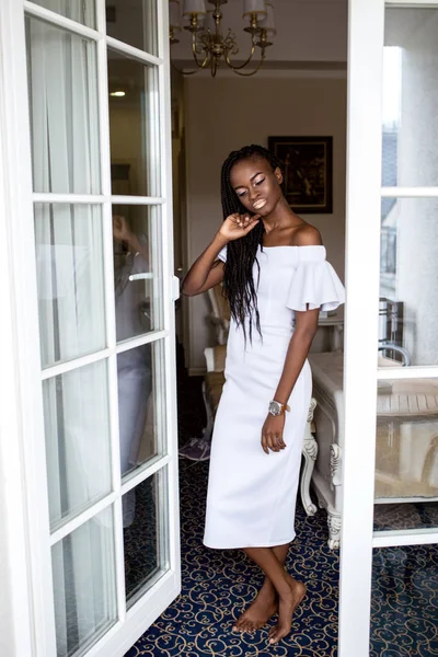 Bella ragazza con la pelle scura e lucida in piedi vicino alla finestra in abito bianco. Donna afro-americana — Foto Stock