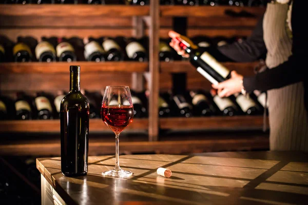 Un verre de vin rouge et une bouteille sur une table. Sommelier tenant une bouteille de vin et debout à côté des étagères avec des bouteilles de vin . — Photo