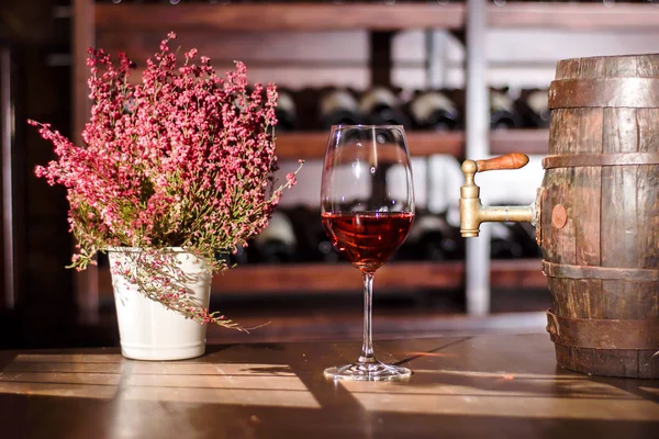 Composizione di vaso di fiori, bicchiere di vino e botte posta su un tavolo di legno. Scaffali con bottiglie di vino su sfondo . — Foto Stock