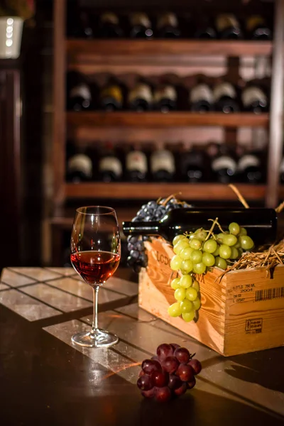 Sammansättningen av vin flaska, glas och box med druvor på ett träbord. Hyllor med vin bootles på en bakgrund. — Stockfoto