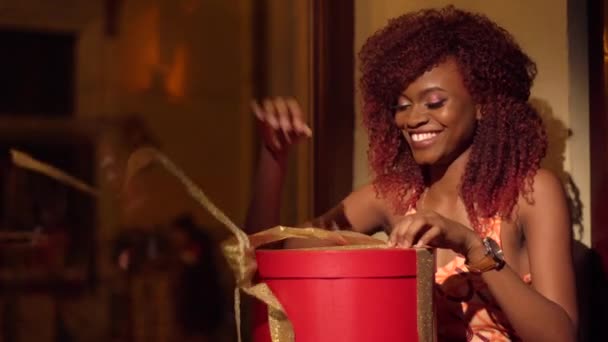 El retrato de mujer afro-americana feliz y sorprendida con el pelo rizado rojo recibiendo la apertura del regalo en caja roja redonda con cintas doradas en la cafetería — Vídeos de Stock