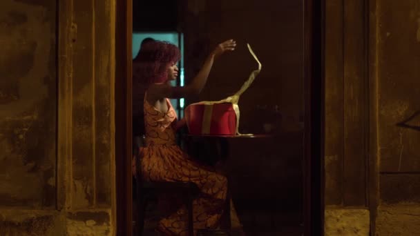 De full-length horizontale foto van de Afro-Amerikaanse meisje met rood krullend haar openen het heden in de ronde rode doos en gouden linten en zitten in de buurt van het venster in het café — Stockvideo