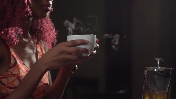 La foto horizontal sensible del primer plano de la mujer afro-americana con el pelo rizado rojo sosteniendo la taza y bebiendo té caliente en la noche en el fondo de la ventana — Vídeo de stock