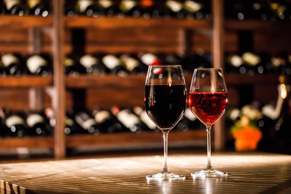 Two glasses filled with wine placed on the wooden table in fromt of the shelves with wine bottles. — Stock Photo, Image