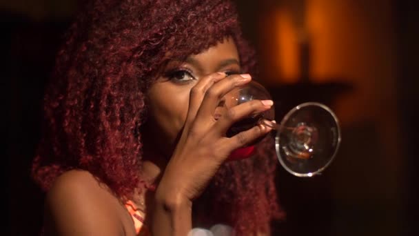Szép közeli portréja az afro-amerikai nő, vörös göndör haja gazdaság ő borospohár, és ivott vörös bort az étteremben — Stock videók