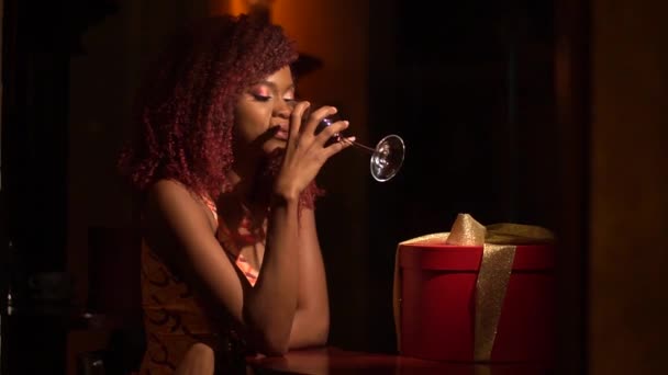 Profilul orizontal al tinerei femei afro-americane care bea vin roșu lângă prezent în cutia rotundă roșie cu panglici aurii, petrecând timp în cafeneaua întunecată — Videoclip de stoc