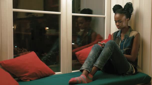 El retrato de la chica afro-americana con coletas africanas charlando con sus amigos, navegando por Internet y sentada en el solft green window-sill rodeado de coloridos cojines . — Vídeos de Stock