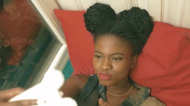 Cep telefonu komik surat ve yeşil yumuşak pencere pervazına kırmızı minder üzerinde yalan gülümseyen Afro-Amerikan kadın kahverengi pigtails selfies, çekici ile yakın çekim portresi