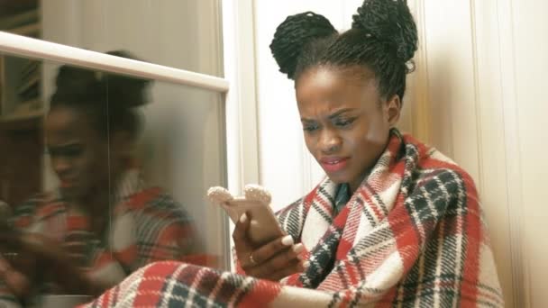 El retrato lateral de la mujer afro-americana ammazada charlando con sus amigos, leyendo, navegando por Internet a través del teléfono móvil mientras está sentada en el anexo ventana-alféizar de la ventana blanca — Vídeo de stock
