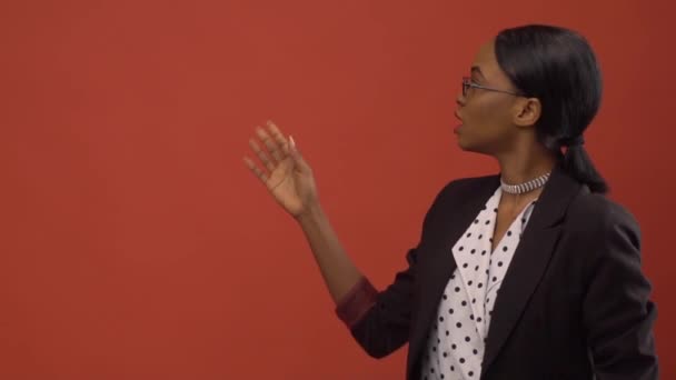 Чорна жінка в куртці піднімає руку, говорячи про щось перед червоною стіною — стокове відео