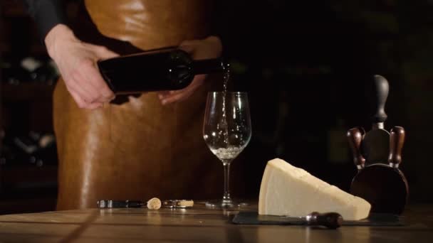 La composizione del formaggio, apribottiglie con tappo di sughero sono disposti sul tavolo di legno. Il sommelier sta riempiendo il bicchiere con il vino bianco nel magazzino del vino . — Video Stock