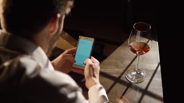 La vista di cui sopra dell'uomo seduto a tavola e chatta, messaggiando tramite il telefono cellulare nella parte anteriore del bicchiere con vino rosso . — Video Stock