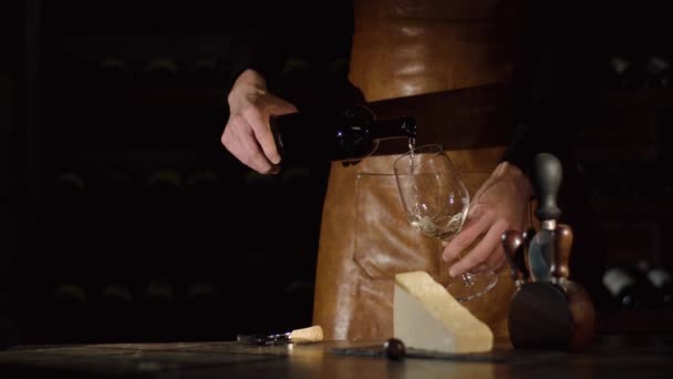 Le mani del sommelier riempiono il bicchiere con il vino bianco e lo mettono sul tavolo di legno vicino al sughero e al formaggio. . — Video Stock