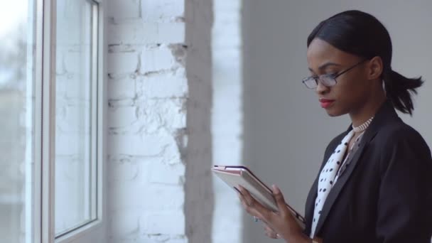 Mujer negra atenta lee algo en su iPad de pie ante una ventana brillante — Vídeo de stock