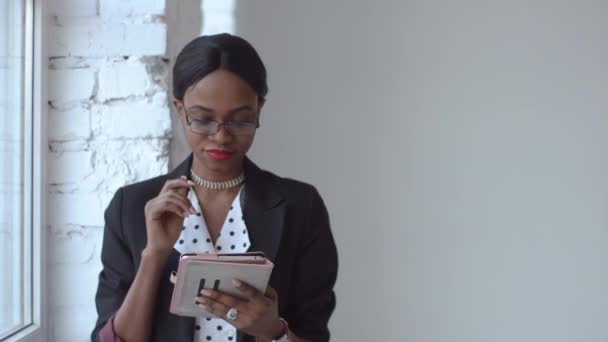 Mulher negra atenta lê algo em seu iPad em pé diante da janela brilhante — Vídeo de Stock