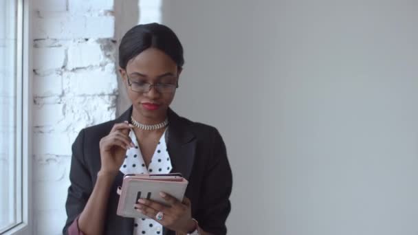 Attentive femme noire lit quelque chose dans son iPad debout devant une fenêtre lumineuse — Video