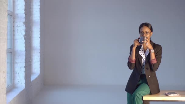 Thoughtful black woman in green trousers drinks coffee sitting on table in office — Stock Video