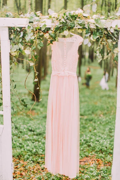 Le dos de la robe rose clair avec de la dentelle suspendue à l'arc blanc décoré de fleurs blanches et placé dans la forêt . — Photo