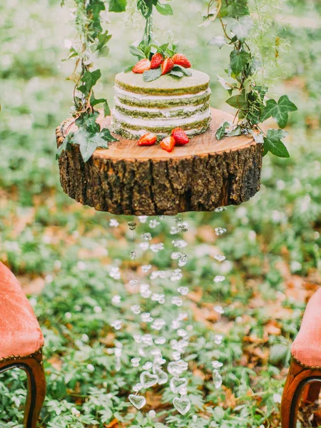 La composición de la torta de esponja verde y blanca con fresas y colocado en el muñón decorado con corazones de vidrio colgante en el bosque . —  Fotos de Stock