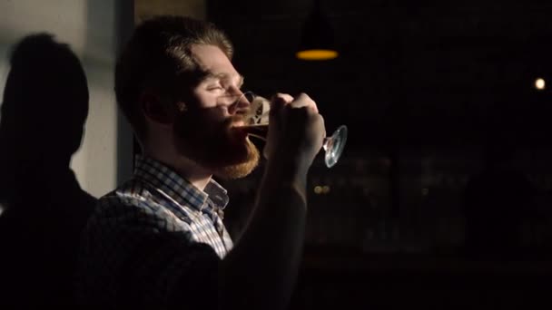 El retrato lateral de cerca del hombre satisfecho bebiendo cerveza y apoyado en la pared en la barra oscura . — Vídeo de stock