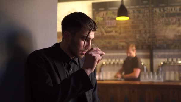 Deprimerad mannen är dricka öl och sortering sitt hår medan bartendern tvår glasögon på den suddig bakgrunden. Den sida ståenden. — Stockvideo