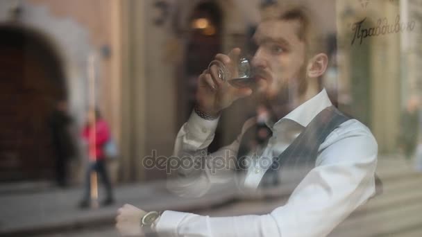 Le portrait horizontal en gros plan à travers la fenêtre du café de l'homme d'affaires élégant assis à la table et buvant du cognac . — Video