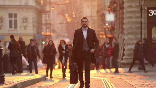 Стильний бізнесмен тримає валізу, перевіряє свої соціальні рахунки через свій мобільний телефон, перевіряє час на руці і ходить у центрі міста під час заходу сонця . — стокове відео