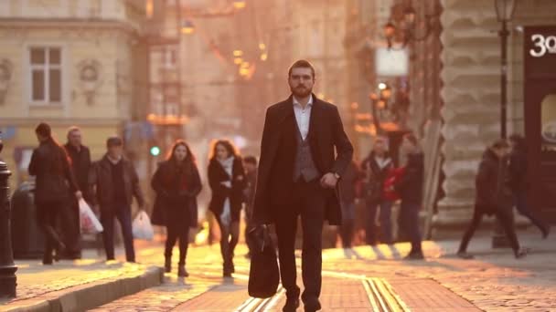 Der stylische Geschäftsmann hält den Koffer in der Hand, checkt per Handy seine Social Accounts, checkt die Uhrzeit auf seiner Handuhr und geht während des Sonnenuntergangs durch die Innenstadt. — Stockvideo