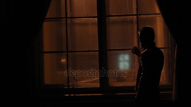 Крупним планом портрет чоловіка, що стоїть, дивлячись через вікно і п'є віскі в темній кімнаті . — стокове відео