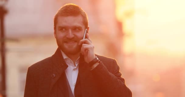 El elegante hombre de negocios sonriente está hablando a través de su teléfono móvil en el fondo del centro de la ciudad durante la puesta de sol brillante . — Vídeo de stock