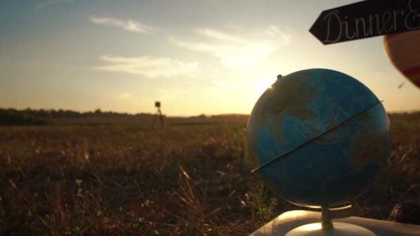 Глобус помещен на винтажный чемодан под деревянной чумой с табличкой в поле во время заката. Крупный план горизонтальной композиции — стоковое видео