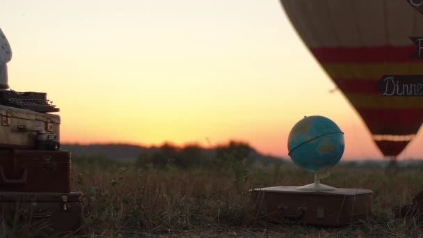 Skład ten kula ziemska umieszczone na brązowy walizki obok rocznika walizki na tle aerostat w polu podczas zachodu słońca. — Wideo stockowe