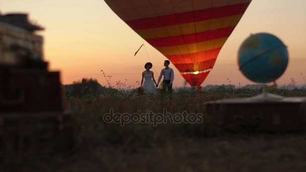 Le globe flou et les valises vintage sont placés à l'avant du couple de marcheurs heureux près de l'aérostat dans le champ pendant le coucher du soleil . — Video