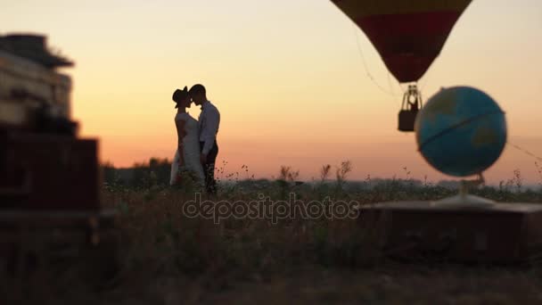O lindo casal vestido vintage está se beijando perto do globo, malas vintage e aerostat voador. A localização do campo durante o pôr do sol . — Vídeo de Stock
