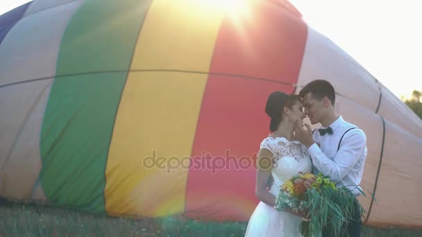 The vintage dressed groom is petting the face of the bride in the black hat and holding the colourful bouquet at the background of the air-balloon. — Stock Video