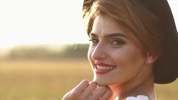 El retrato de cerca de la mujer con lápiz labial rojo, cateyes y sombrero negro sonriendo a la cámara al fondo del campo soleado . — Vídeos de Stock