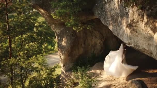 De mooie bruid met de sluier is spelen met de lange witte trouwjurk en ronddraaien op de rots in de groene zonneveld. — Stockvideo