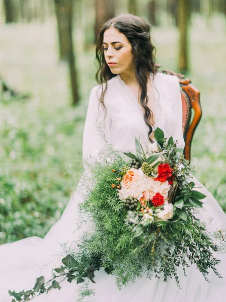El hermoso retrato de la mujer en el vestido de novia largo y blanco sosteniendo el enorme ramo verde con flores y sentado en el chait en el bosque verde . — Foto de Stock