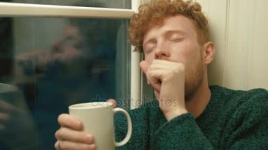 Sıcak çay içme öksürük ve pencere yakın oturan hasta adam altın kıvırcık saçlı beyaz Kupası tutarak. Yan yakın çekim portre