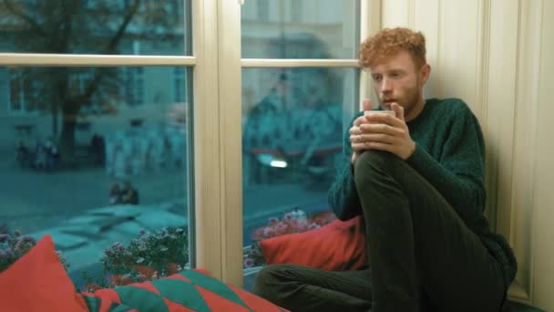 Il ritratto laterale del ragazzo biondo riccio circondato da cuscini che bevono tè caldo e si siedono sul davanzale della finestra. Ha il raffreddore. . — Video Stock