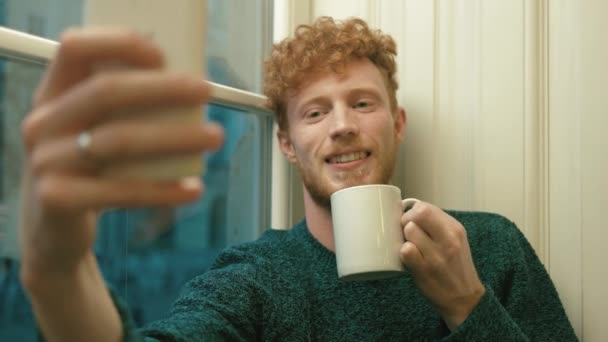 Portret mężczyzny z blond włosy kręcone robienia selfie uśmiechający się i poważne, przy filiżance gorącej herbaty — Wideo stockowe