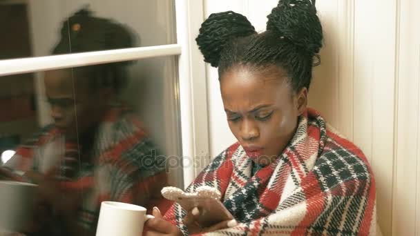The horizontal close-up portrait of the surprised afro-american woman with african pigtails, reading articles via mobile phone and drinking tea under the red knitted plaid near the window. — Stock Video