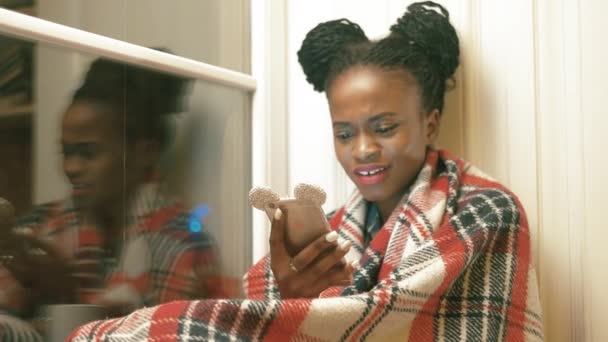 Sorrindo menina afro-americana com tranças africanas marrom está lendo o artigo engraçado, conversando com seus amigos e navegando na Internet através de seu telefone celular, envolto no xadrez de malha vermelha — Vídeo de Stock