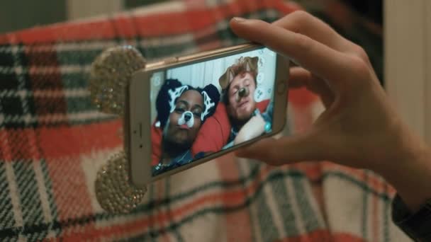 The photo of multi-race couple taking selfies using snapchat app via mobile phone while lying under the red knitted plaid. The mask of brown dog and dalmatian. — Stock Video