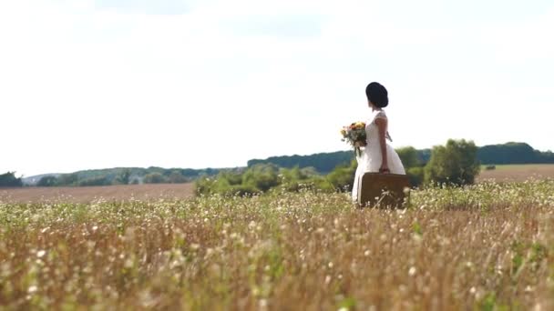 Боковой вид женщины в белом платье и черной шляпе, держащей огромный букет полевых цветов и несущей старинный чемодан на фоне солнечного поля . — стоковое видео