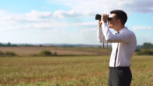 The smiling groom in the vintage suit is watching at the sky using binoculars at the background of the sunny field. — Stock Video